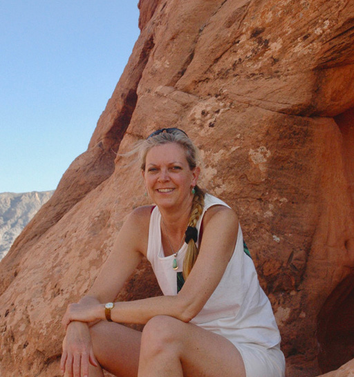 The Author, Debbi, sits in light summer clothing on a ledge against the steep red rock wall of a shaded slot canyon in Grand Staircase-Escalante National Monument, Utah. She smiles and leans slightly forward with her elbows propped on her knees.